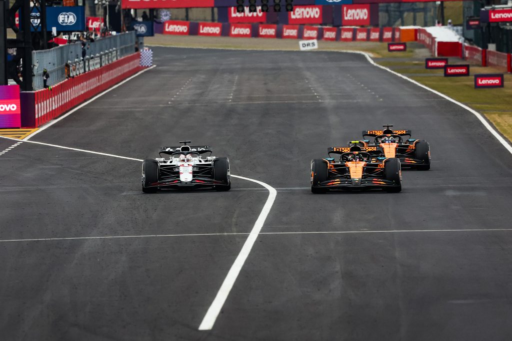 Max Verstappen’s Red Bull #1 (dark blue car) leads down the straight, with Lando Norris’s McLaren #4 (orange car) chasing to the right and Oscar Piastri’s McLaren #81 (orange car) following just behind his teammate.