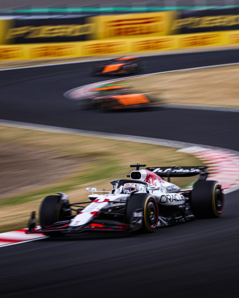 Max Verstappen’s Red Bull (dark blue car) #1 attacks the kerb in the foreground, while two McLaren (orange cars) blur through the next section of the Suzuka esses ahead.