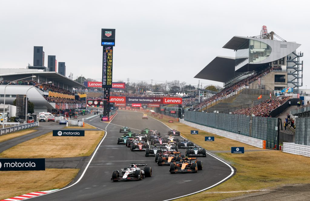 The lead Racing Bulls (blue car with white and silver highlights) heads the pack into the opening sector, with two McLaren (orange cars), Norris’s #4 and Piastri’s #81, running side by side in pursuit while a Mercedes (silver car with black element...